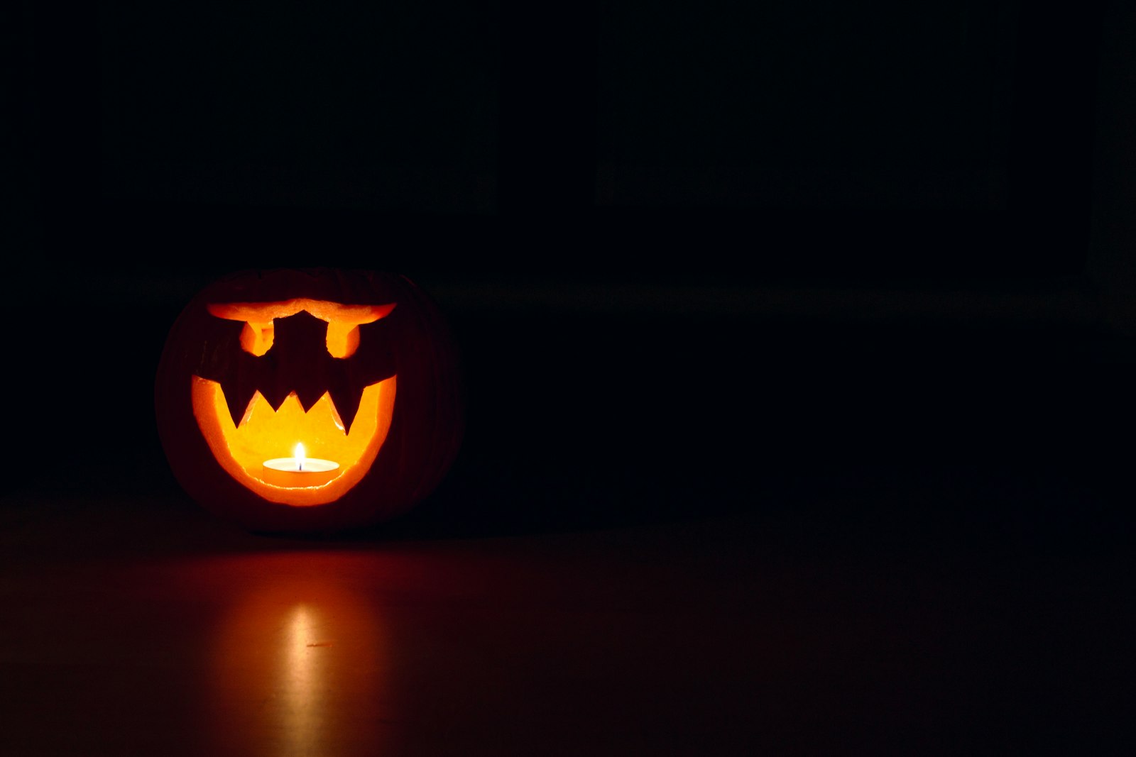 A single carved jack-o'-lantern glows orange at the left of an almost black frame, its light pooling on the dark surface below.