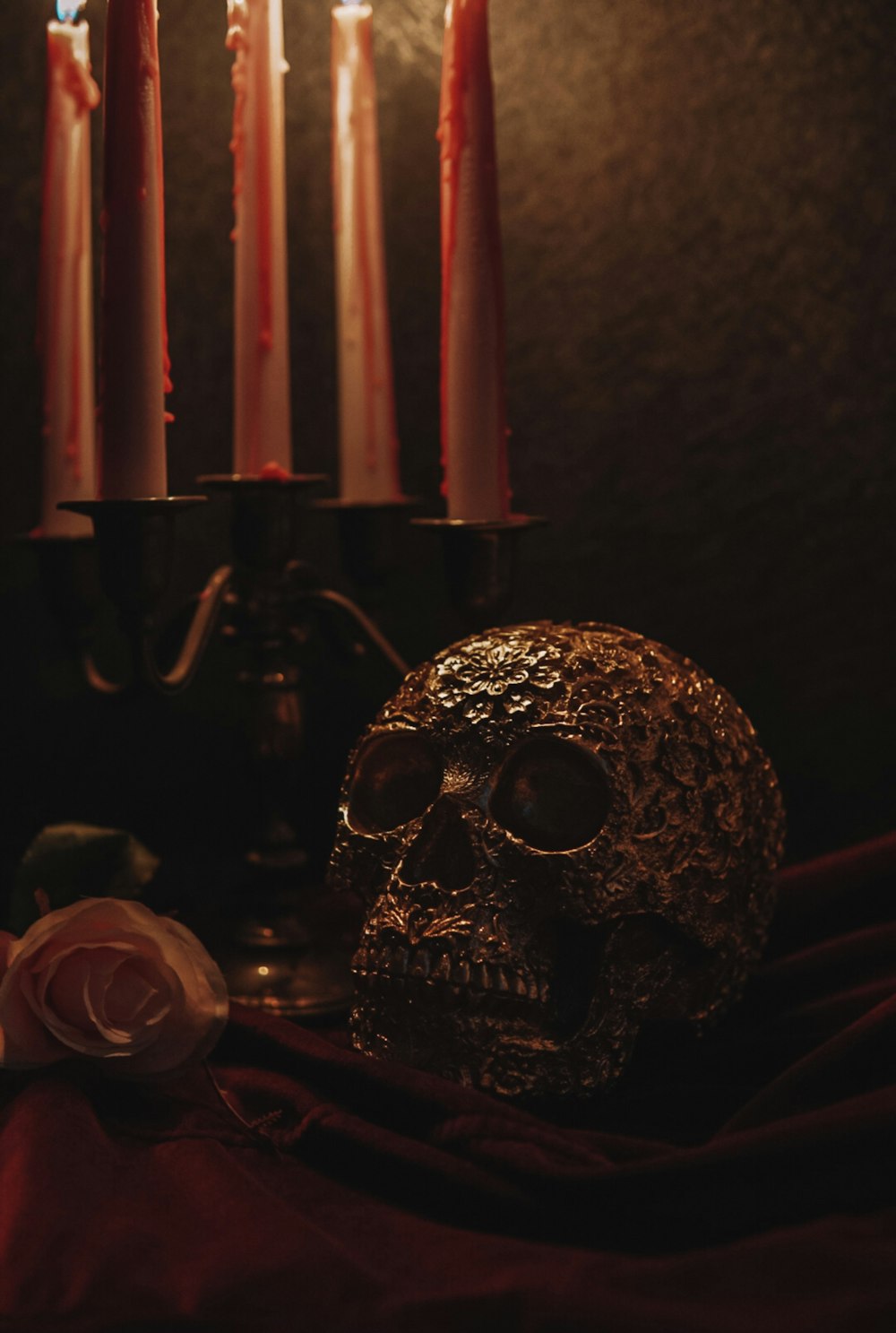 Tall cream taper candles streaked with red wax drips in a dark candelabra, beside an ornate bronze-gold skull and a pale rose on dark red fabric.