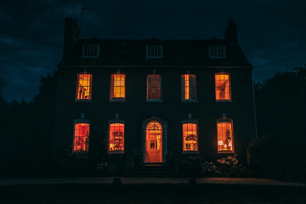 A symmetrical brick Georgian country house at night, most of its windows and the front door glowing orange under a dark, cloudy navy sky.