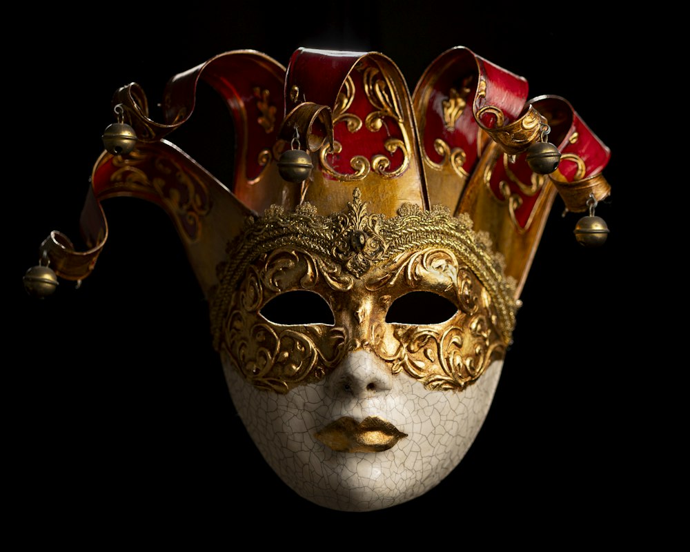 A white crackle-glaze Venetian jester mask with gold filigree, a red and gold peaked cap and small bells, lit against a black background.