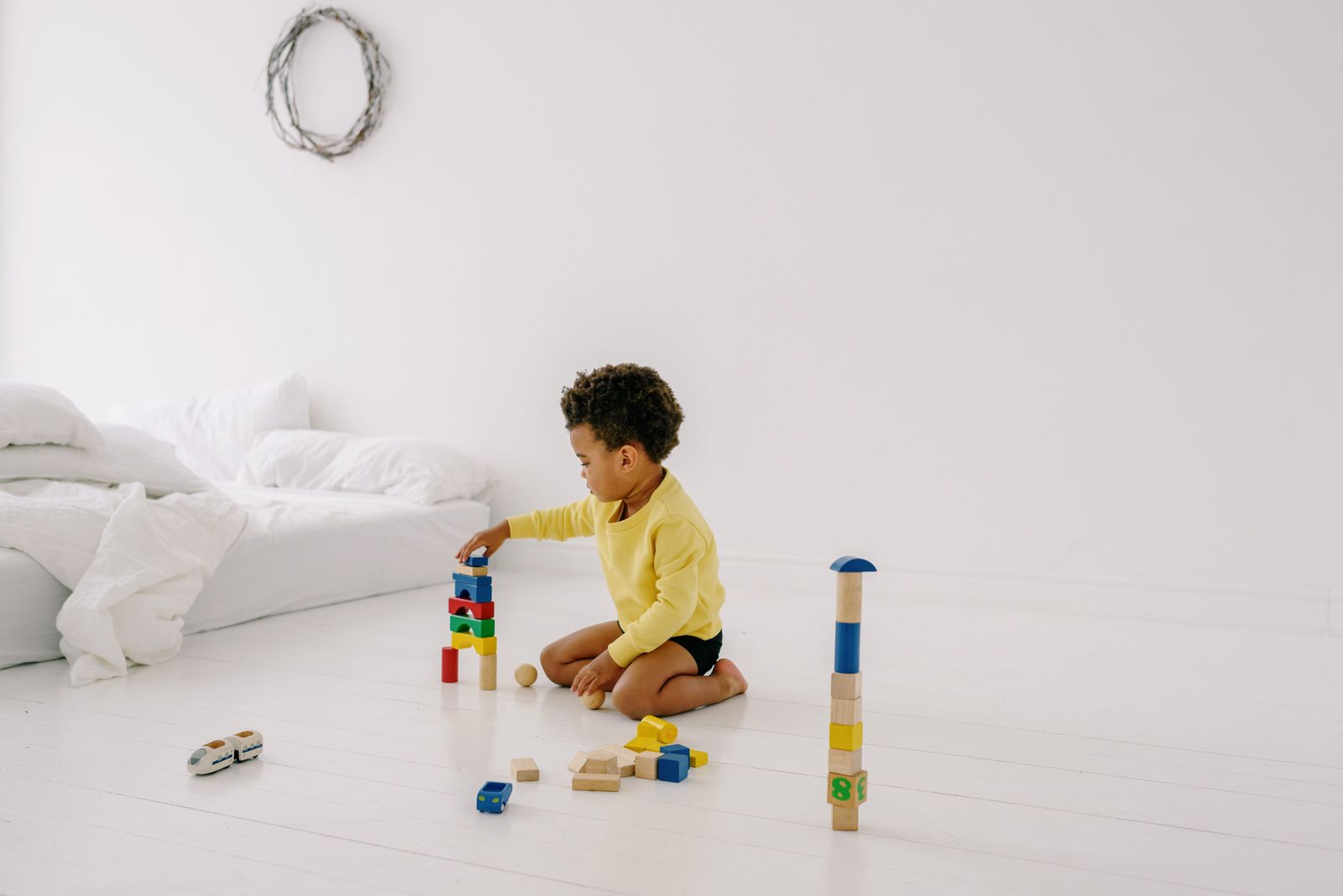 A small child in a yellow top kneels on a white bedroom floor, building a tower from colorful wooden blocks with a few toys scattered around.