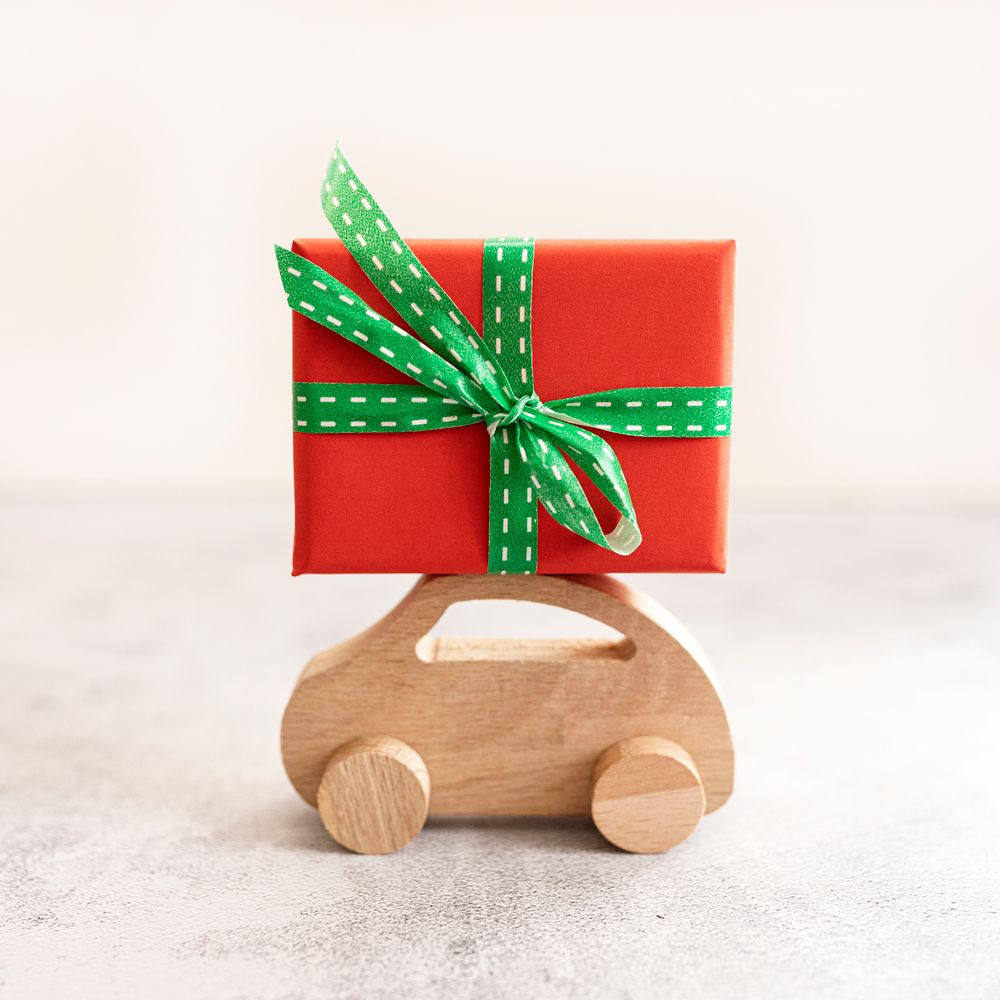 A wooden toy car carries a red gift box tied with a green dotted ribbon on a pale concrete surface.