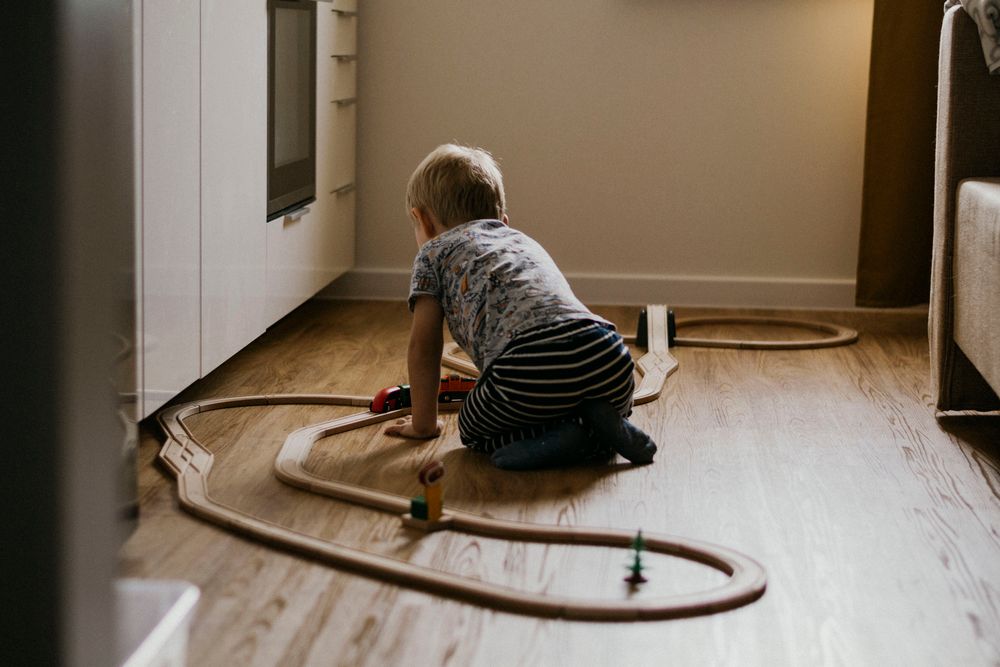 A toddler seen from behind kneels on a wooden floor, playing with a looping wooden train track beside a bedroom wall.