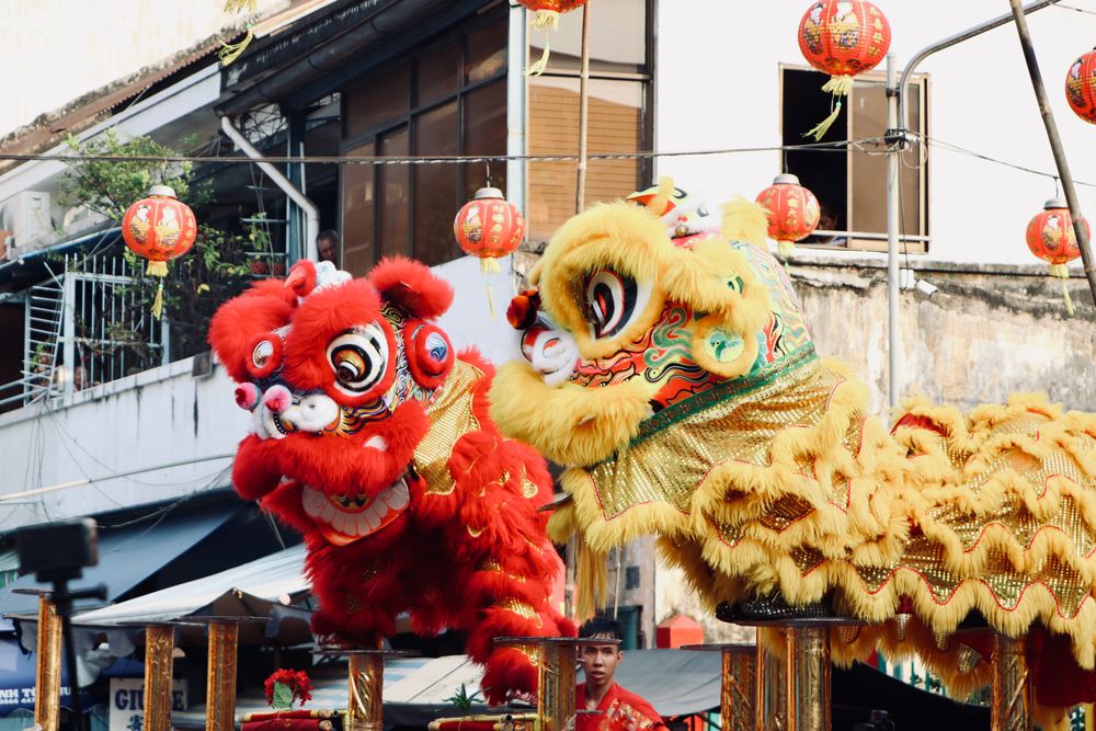 Lion dance performers in red and yellow costumes at a festival with lanterns behind.