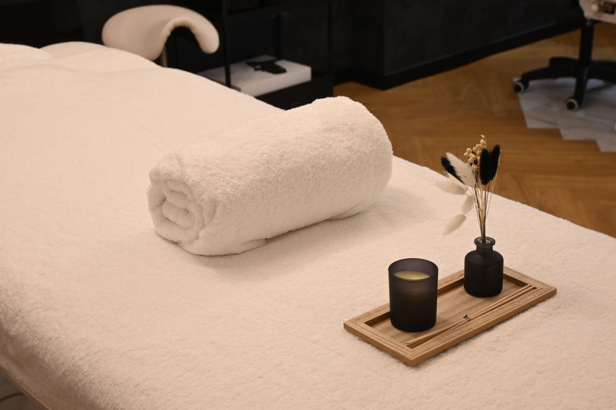 A spa treatment table covered in white towelling with a rolled white towel and a small wooden tray holding a dark candle and a reed diffuser.