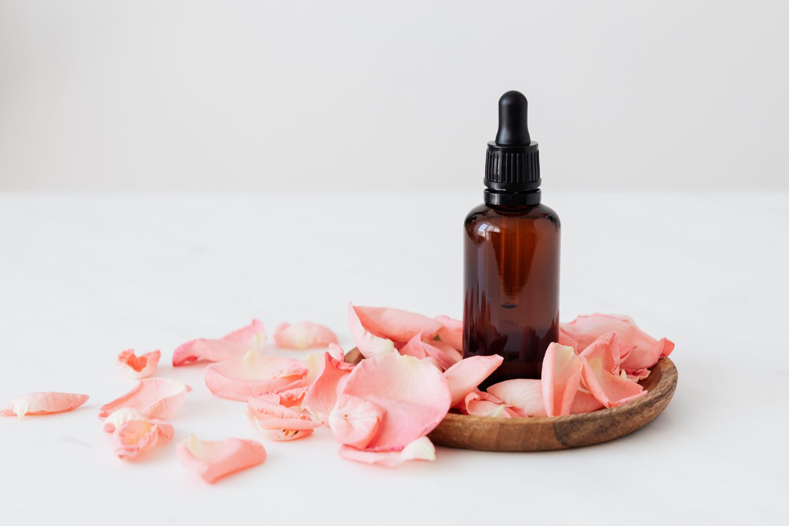 An amber glass dropper bottle with a black cap stands on a small round wooden plate among pale pink rose petals on a white marble surface.