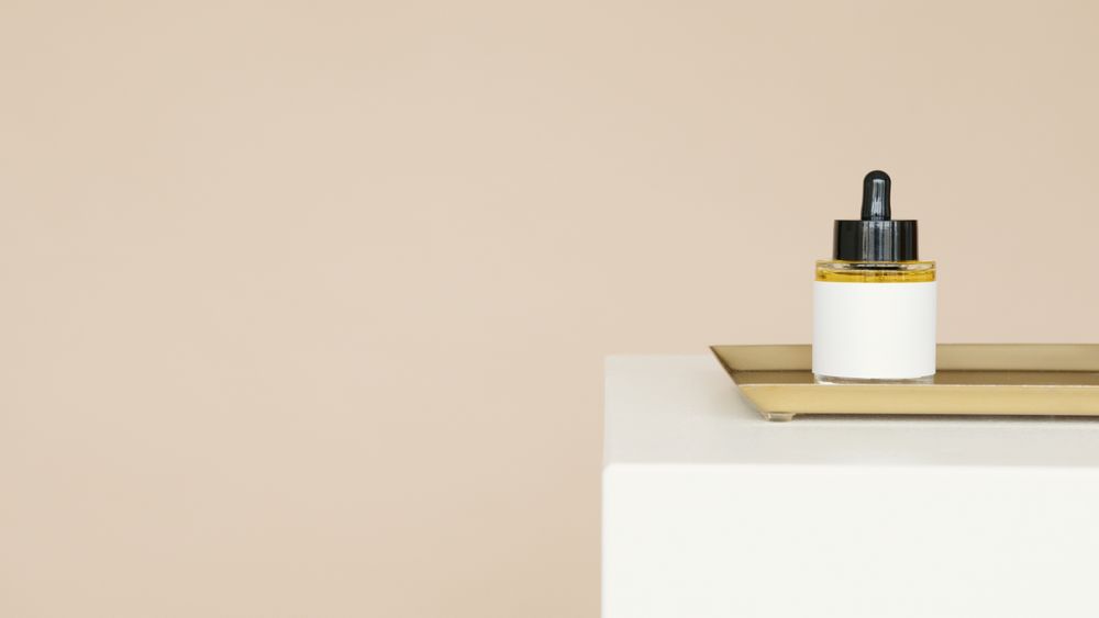 A small white dropper bottle with a black cap sits on a white block with a thin gold tray against a flat warm beige backdrop.