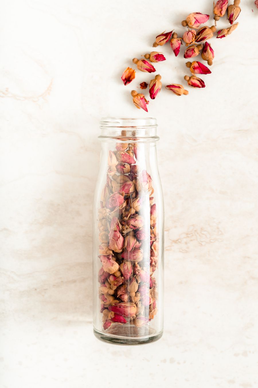 From above, a clear glass bottle packed with dried red rosebuds on pale veined marble, with more buds scattered from its neck.