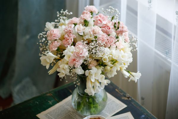 A bouquet of pink carnations, white freesia and baby's breath in a glass vase on a weathered green table