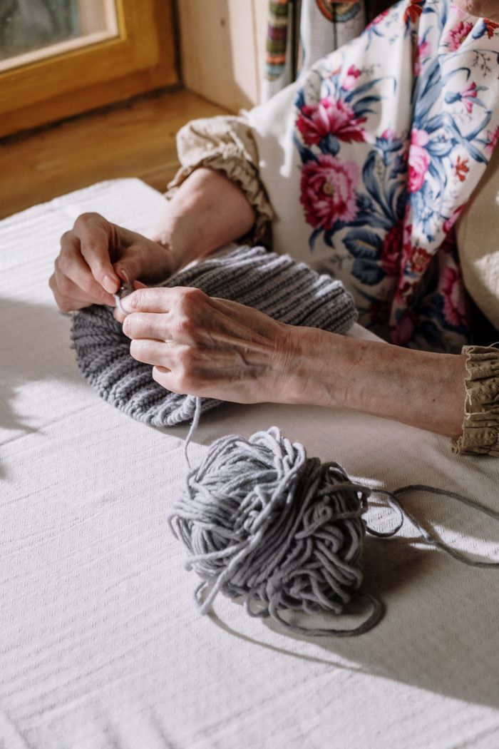 An older woman's hands knitting a grey ribbed piece beside a ball of grey yarn