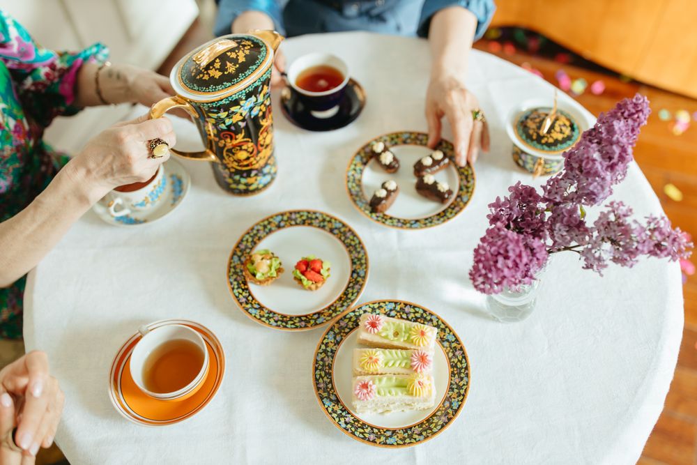 Overhead view of a round white table set with gold-rimmed floral china, tea cups, small cakes and a vase of purple lilac while hands pour tea