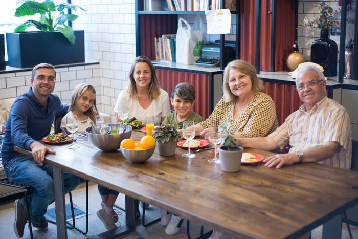 Three generations of a family smiling at one long wooden table laden with bowls of salad and fruit