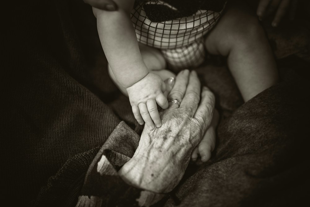In a dark sepia photograph, a baby's tiny hand rests on an older woman's wrinkled, ringed hand
