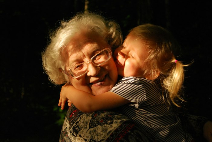 An elderly woman with white hair and glasses hugged and kissed on the cheek by a young child, in a warm golden glow