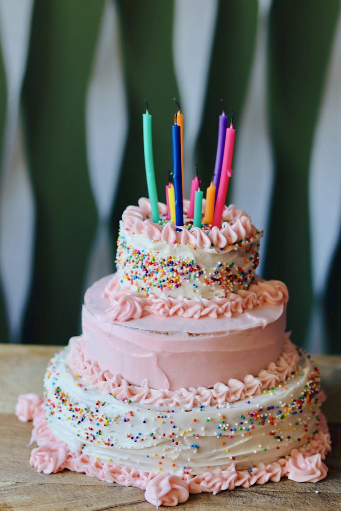 A small pink tiered birthday cake with piped frosting, sprinkles and tall unlit candles on a wooden board