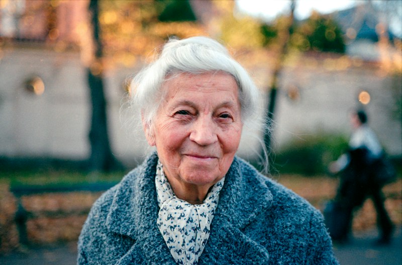 A serene elderly woman with white hair, a blue-grey wool coat and a patterned scarf, smiling gently against golden autumn light