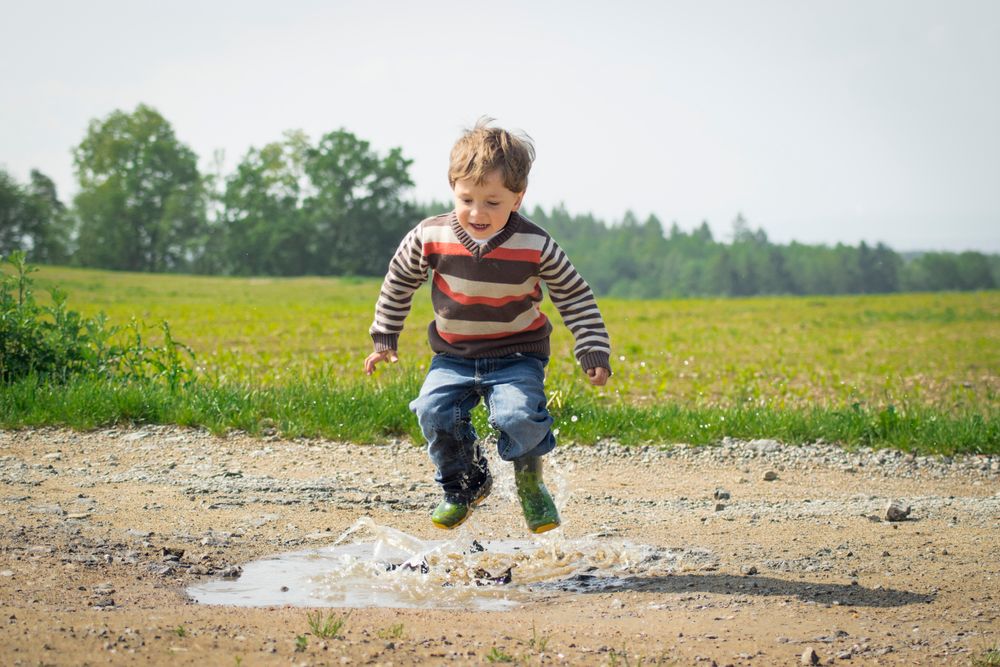A boy in a striped sweater and green wellies jumps over a small puddle on a gravel path in front of a green meadow and a pale sky.