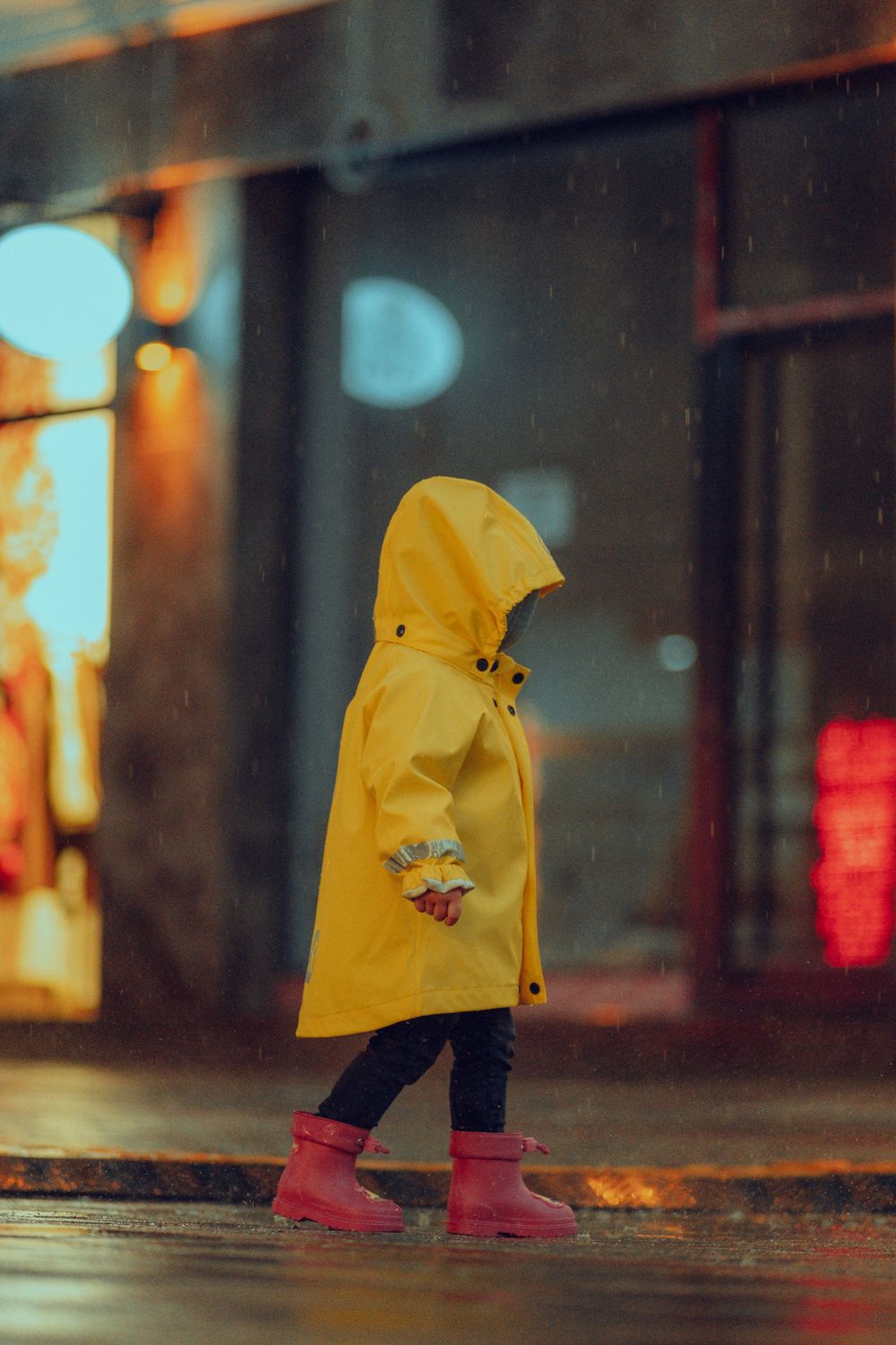 A small child in a yellow hooded raincoat with gathered reflective cuffs and pink rubber boots walks along a wet street in the rain, seen in profile with the hood up.