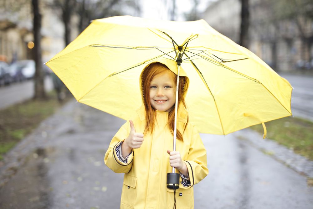 A smiling red-haired girl in a yellow raincoat holds a bright yellow umbrella and gives a thumbs up on a wet, tree-lined street on a grey rainy day.