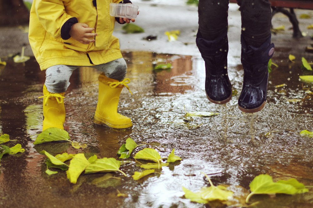 A child in a yellow raincoat and yellow rubber boots crouches in a puddle scattered with green leaves while a second child in dark boots jumps beside them.