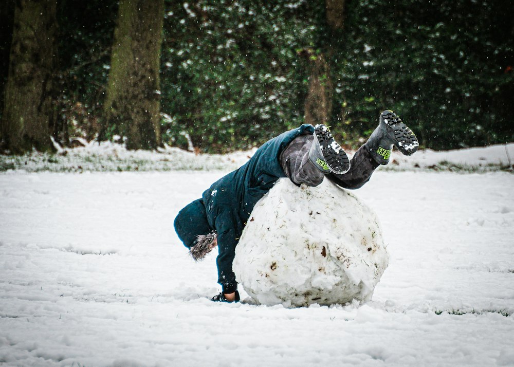 A child in a dark teal jacket tumbles head-down over a large snowball in a snowy park with dark hedges behind.