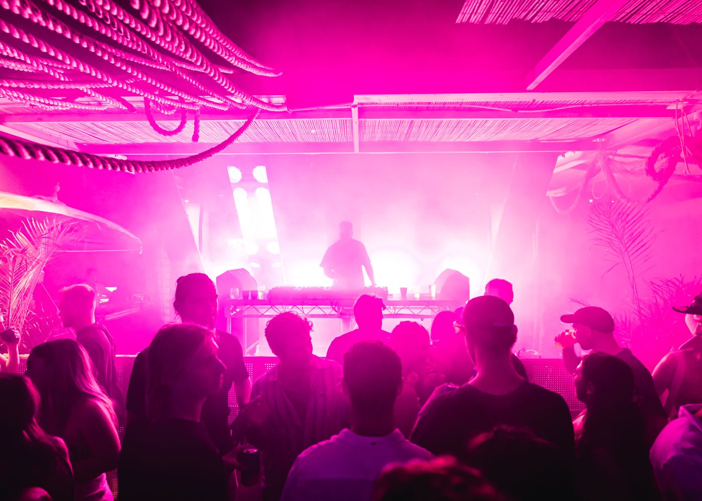A crowd seen from behind dancing in front of a DJ booth, with the DJ in silhouette and the whole room washed in hot-pink light and haze.