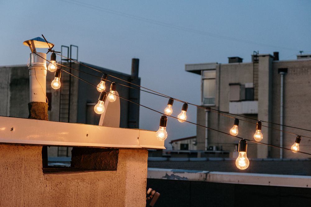 A string of warm filament bulbs stretched across a rooftop at dusk, with a blue twilight sky and neighbouring apartment buildings behind.