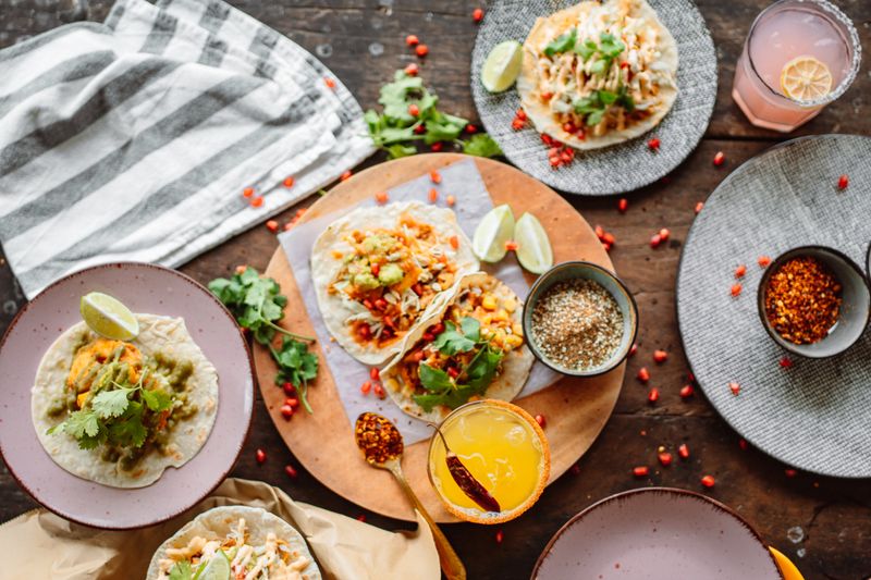 Top-down view of tacos on plates and a wooden board with lime wedges, cilantro, chilli flakes, pomegranate seeds and an orange drink on a dark rustic table.