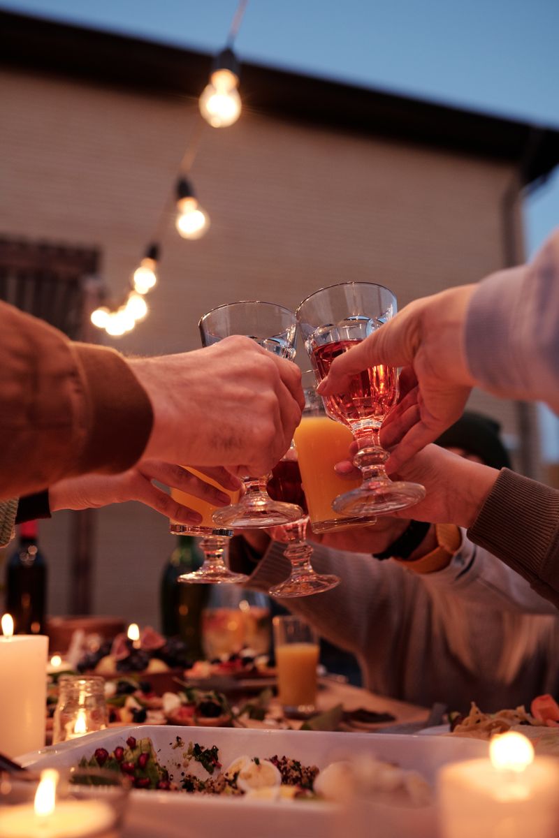 Friends' hands clinking wine glasses and orange juice over a candlelit table at dusk, with warm filament bulbs strung overhead against a blue evening sky.