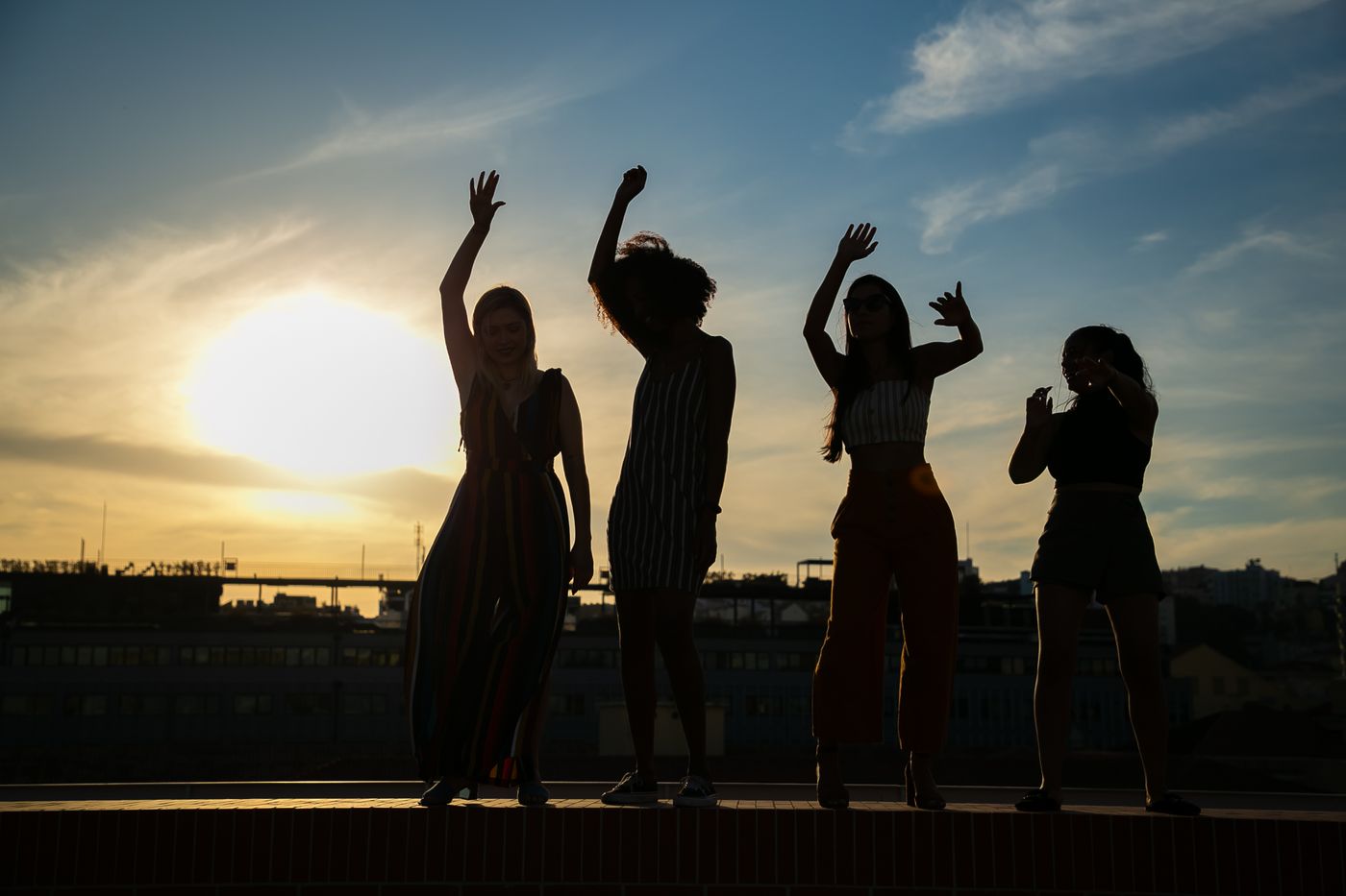 Silhouettes of four women dancing with raised arms on a rooftop ledge against a glowing orange sunset with wispy clouds.