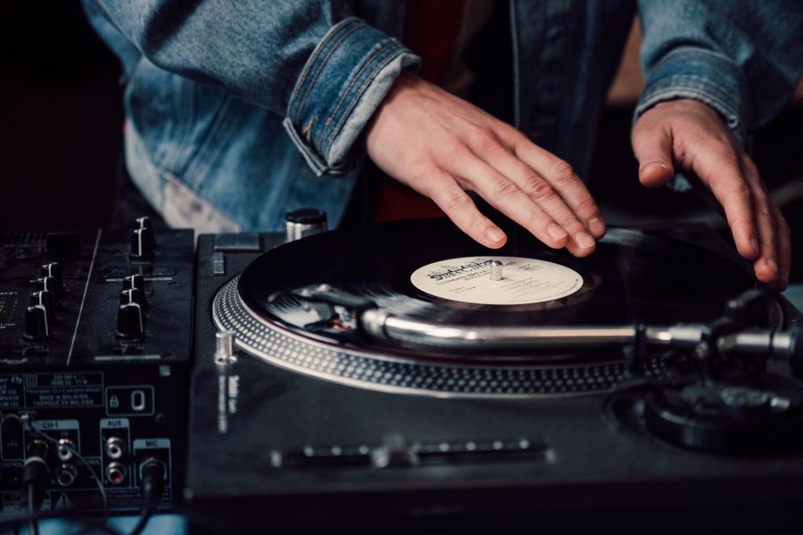 A DJ in a denim jacket resting both hands on a spinning vinyl record on a turntable, next to a mixer with knobs and faders.