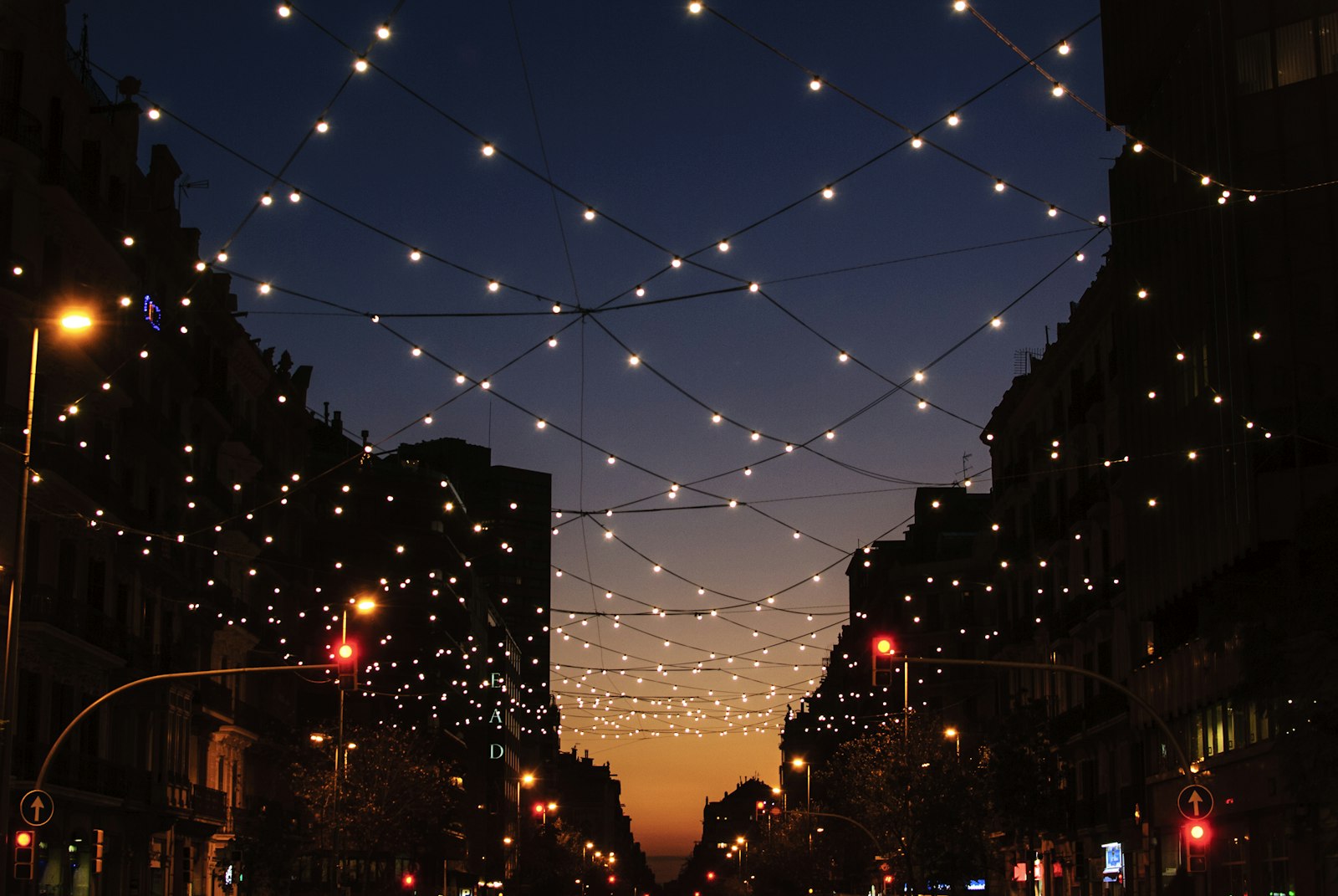 Strings of small white festoon lights crisscrossing a city street at dusk under a deep navy sky with an orange glow on the horizon.