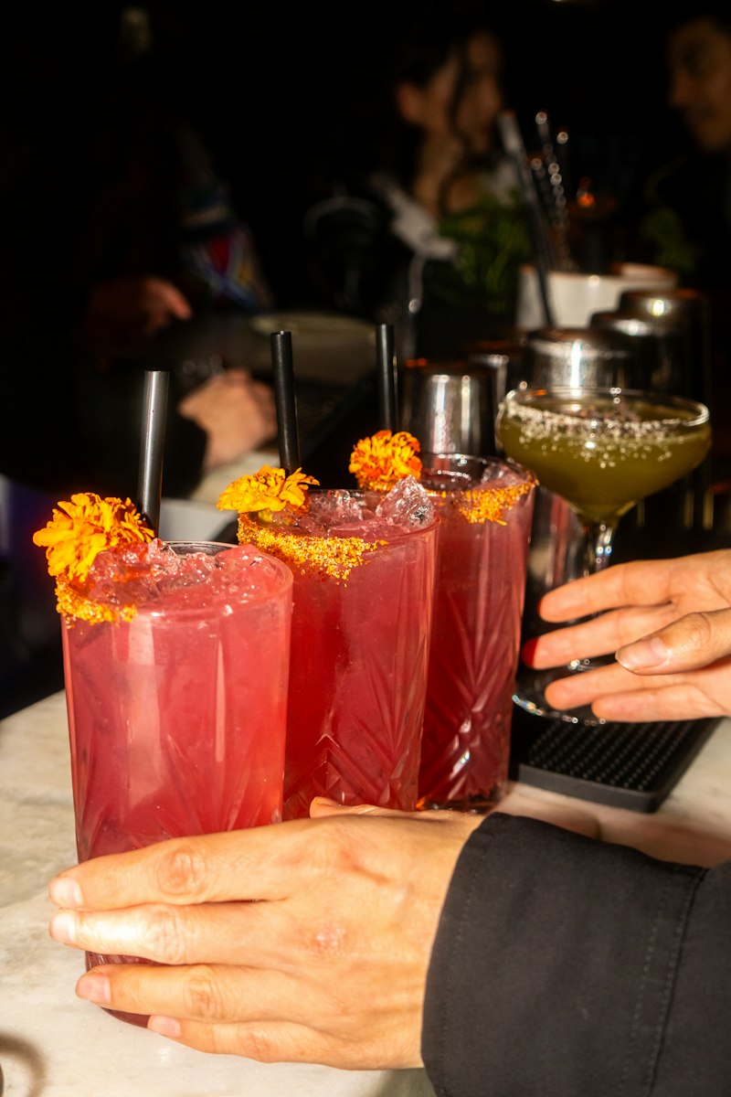 Three crimson cocktails garnished with orange marigold flowers on a pale bar top at night, with hands setting them down and a green cocktail beside them.