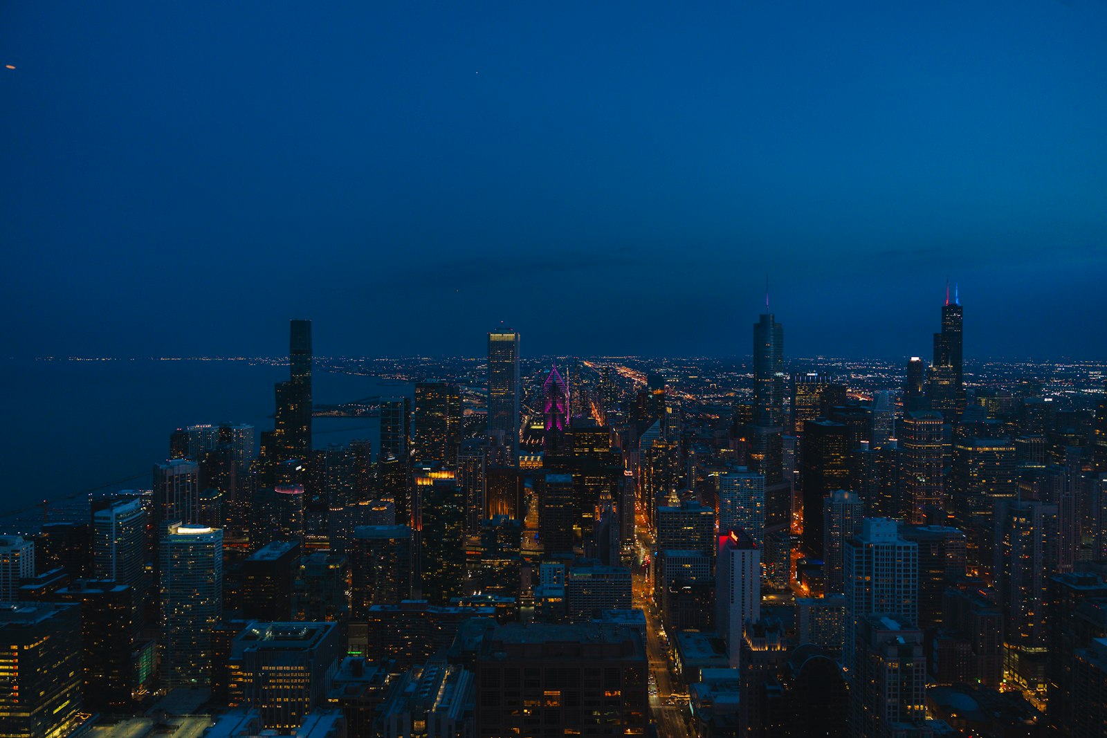 A high-rise city skyline seen from above at night, with lit towers, glowing streets and the lake edge under a deep blue dusk sky.