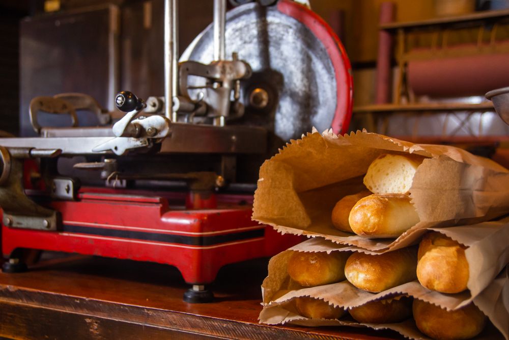 Fresh crusty rolls tucked into brown paper bags on a wooden counter beside a vintage red-and-steel slicer.