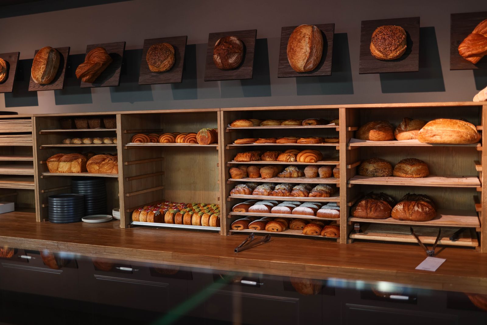 A long wooden bakery counter in front of shelves of fresh loaves, rolls and pastries, with round loaves mounted on small plaques along the wall above.
