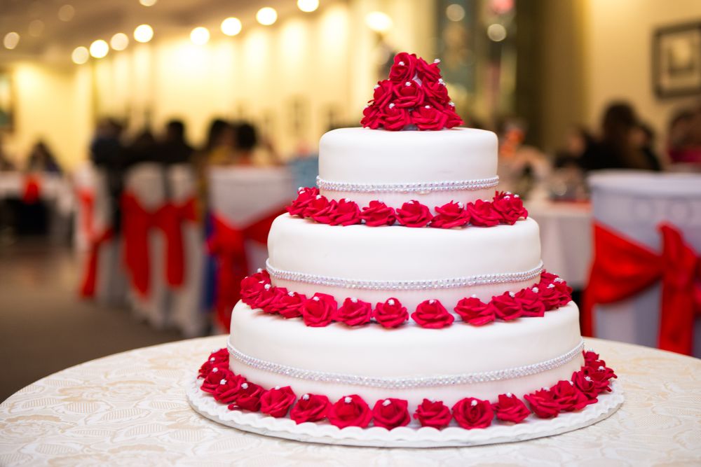 A three-tier white wedding cake ringed with red sugar roses and silver beading, with a cluster of roses on top, on a lace-covered table in a warmly lit banquet hall.