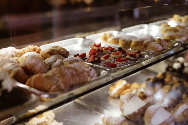 Low view through a glass bakery case at silver trays of powdered-sugar puff pastries, strawberry and berry tartlets and cream-filled buns.