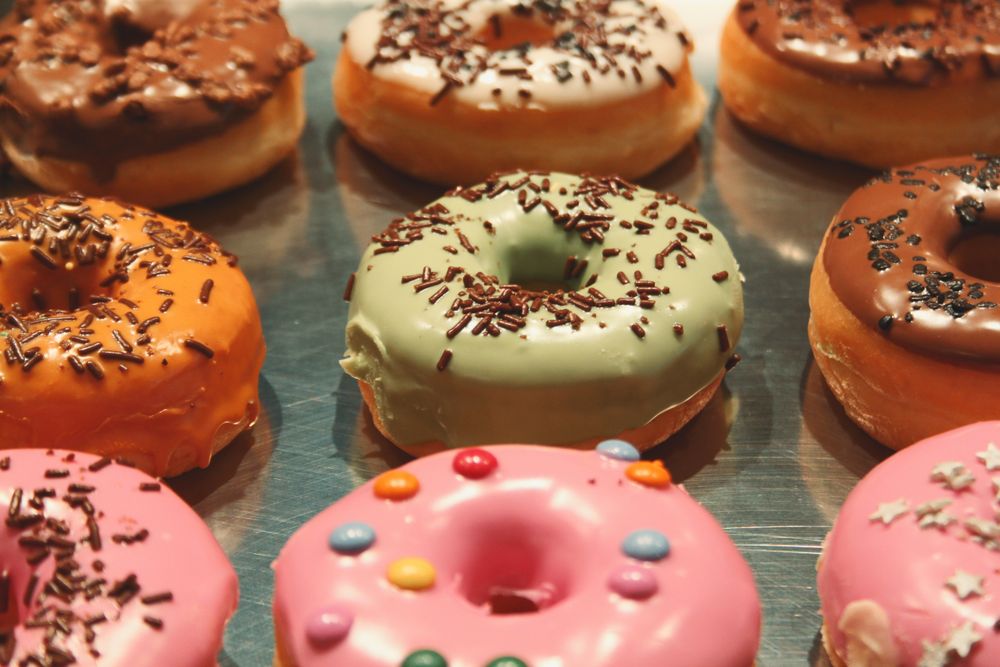 Rows of iced donuts with chocolate sprinkles in pink, pistachio-green and chocolate glazes on a metal tray.