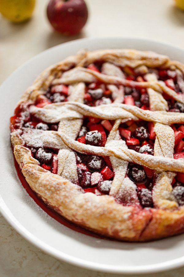 A lattice-topped fruit pie of dark cherries and diced apple dusted with powdered sugar on a white plate.