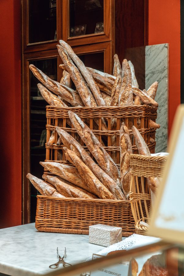 Tall wicker baskets stacked with crusty baguettes beside a burnt-orange wall and a marble counter in the bakery.