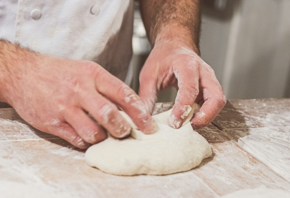 A baker in a white jacket shapes a round of dough with flour-dusted hands on a floured wooden table.