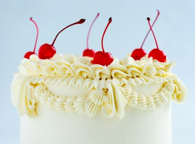 A tall white buttercream layer cake with piped ruffles around the top and glossy red cherries on top, against a pale blue background.