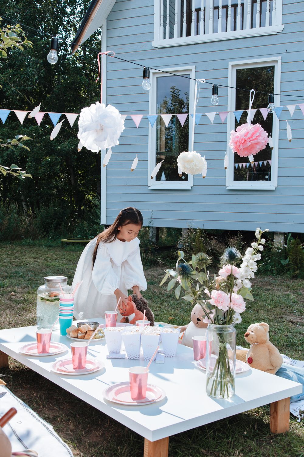 A girl in a white dress arranges a pastel garden party table with flowers, teddy bears, pink cups, paper pom-poms and bunting in front of a pale blue wooden house.