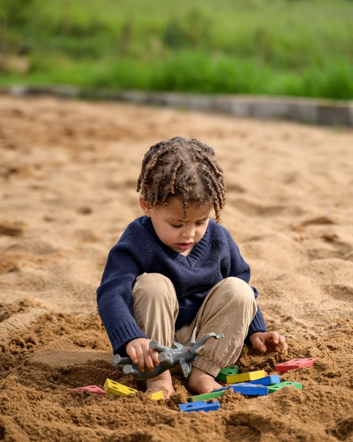 A toddler with twisted braids sits barefoot in warm sand holding a toy dinosaur among colorful wooden shape-sorter pieces.
