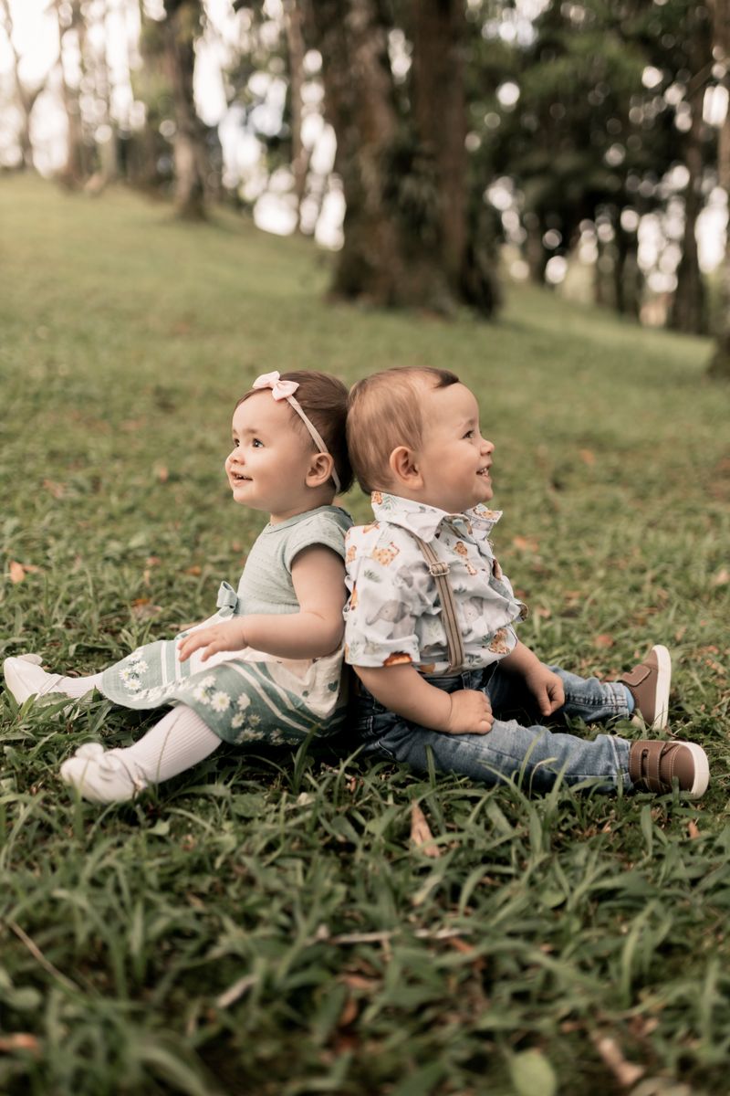 Two babies in soft green and pale printed outfits sit back to back on a grassy slope under tall trees, both smiling and looking off to the side.