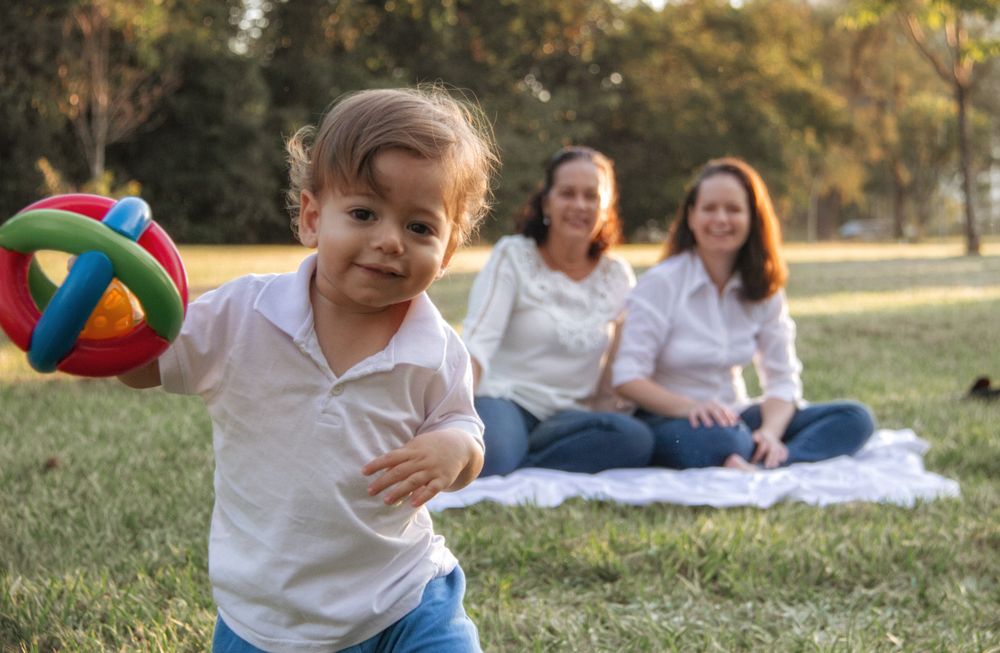 A toddler in a white polo shirt toddles toward the camera holding a colorful ring toy while two smiling women sit chatting on a white blanket in a sunlit park behind him.