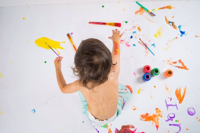 Overhead view of a toddler reaching out to paint on a white floor splashed with bright paints, brushes and paint pots.
