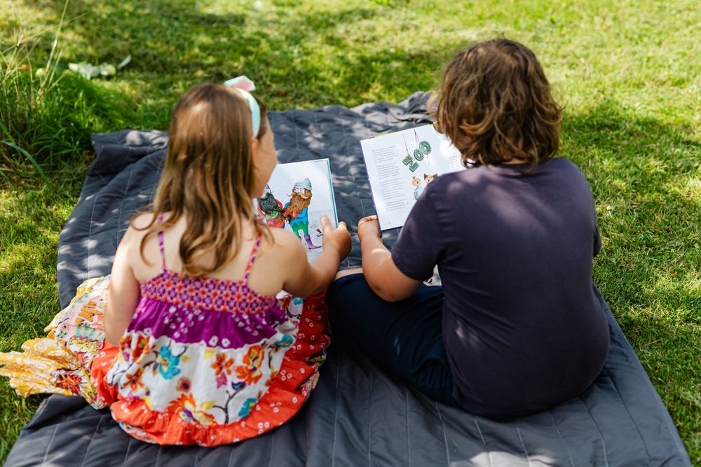 Two children seen from behind sit side by side on a gray quilted blanket on sunlit grass, each holding an open picture book.