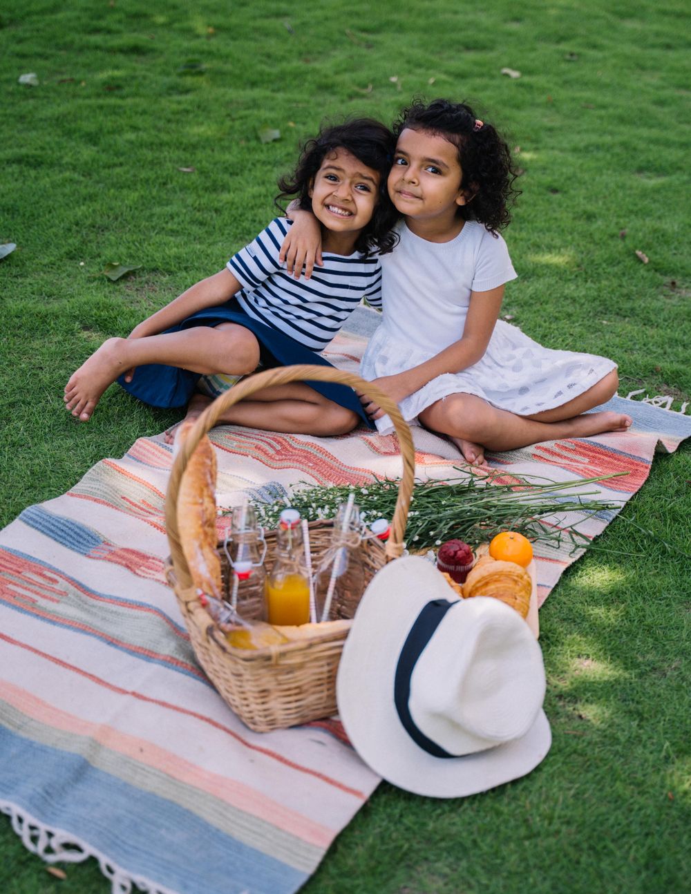 Two smiling girls sit with their arms around each other on a pastel striped picnic blanket beside a wicker basket of snacks, with a white sun hat on the lawn.